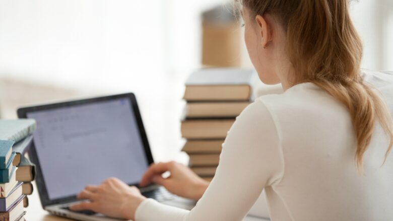 A student reviews a final essay draft on a laptop beside stacks of books