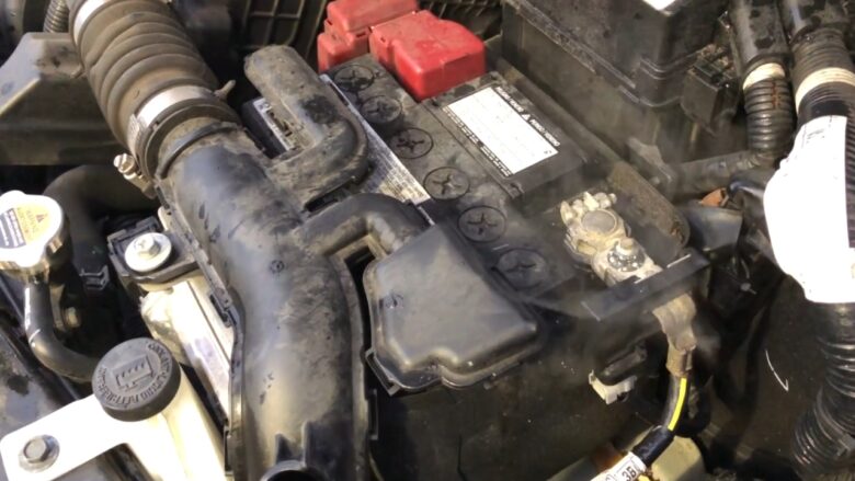 A dusty car battery sits in an engine bay with visible terminals and cables