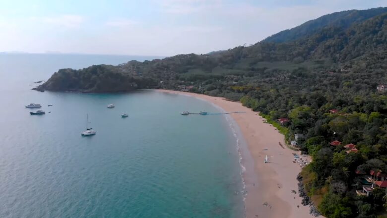 Aerial view of a quiet Koh Lanta beach with calm water, boats and forested hills