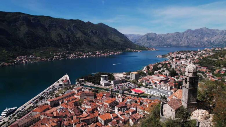 Aerial view of Kotor's red-roofed Old Town, stone church tower and surrounding mountains.