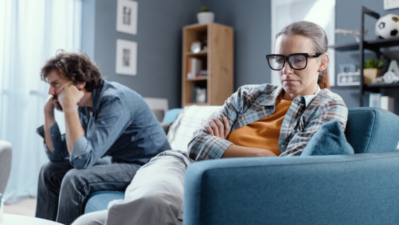 A tense couple sits apart on a sofa at home.