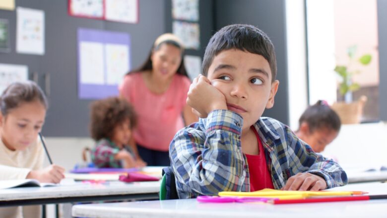 A boy sits at his desk in class as a teacher talks behind him