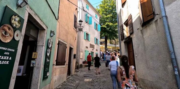 Tourists walk along a cobblestone street past pastel houses with wooden shutters in Istria.