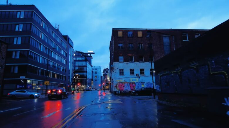 Rainy Manchester street at dusk with traffic and graffiti-covered walls.