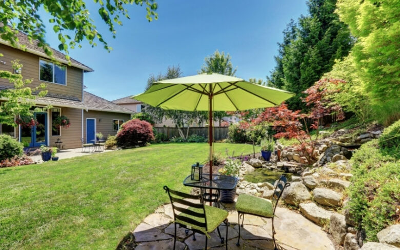 Dream backyard with a green lawn, stone patio, outdoor seating, shade umbrella, and lush landscaping around a family home.
